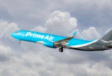 A light-blue Amazon Prime Air cargo jet flies takes off through the clouds.